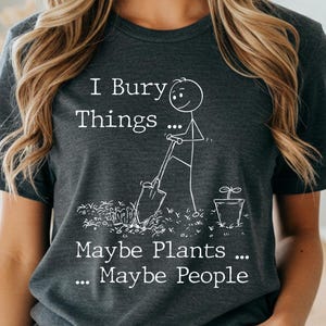 Op de afbeelding: Donkergrijs gemêleerd T-shirt met de tekst "I Bury Things... Maybe Plants... Maybe People" en een stokfiguur met een schop. Het shirt heeft ook de woorden "Dark Heather Gray" aan de onderkant.
