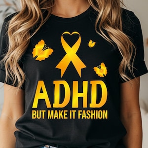 Könnte beinhalten: Ein schwarzes Kurzarm-T-Shirt mit einer gelben Schleife in Herzform, drei gelben Schmetterlingen und dem gelben Schriftzug "ADHD BUT MAKE IT FASHION" auf der Vorderseite. Das Design ist mittig auf der Brust des Kleidungsstücks platziert.