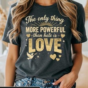 Puede incluir: Camiseta gris jaspeado oscuro con el texto dorado "The only thing more powerful than hate is LOVE". El diseño incluye corazones, una paloma y un sol.
