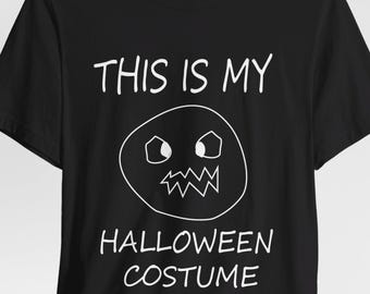 May include: A black short-sleeved t-shirt featuring white text that reads THIS IS MY HALLOWEEN COSTUME. Centered between the lines of text is a simple white line drawing of a circular face with jagged teeth and angular eyes.
