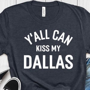 Op de afbeelding: Een heather navy t-shirt met korte mouwen en de witte tekst Y'ALL CAN KISS MY DALLAS op de voorzijde. Het shirt heeft een ronde hals en een standaard pasvorm, afgebeeld naast zwart-witte sneakers en een blauwe spijkerbroekshort.