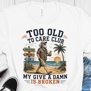 May include: A white t-shirt featuring a graphic of a person with a beard and sunglasses wearing a floral shirt and shorts, walking on a beach with a palm tree and sunset. The text reads TOO OLD TO CARE CLUB, VACATION MODE ON, MY GIVE A DAMN IS BROKEN.
