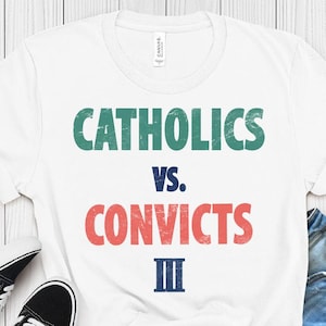 Peut inclure: T-shirt blanc avec l'inscription "CATHOLICS vs. CONVICTS III" en vert, rouge et bleu. Le t-shirt est associé à des baskets noires et blanches et à un short en jean, pour un look décontracté.