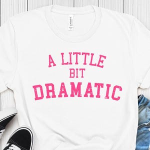 May include: White t-shirt with the text "A LITTLE BIT DRAMATIC" in pink, paired with black and white trainers and denim shorts. The shirt is knotted at the waist. The background is a light wood surface.
