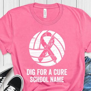 以下が含まれることがあります： 白いバレーボールのデザインとピンクのリボンが付いたピンクのTシャツ。「DIG FOR A CURE SCHOOL NAME」の文字が下にプリントされています。デニムショーツと黒いスニーカーと合わせています。