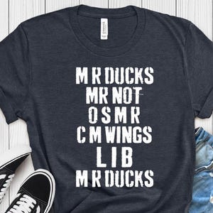 May include: Heather navy t-shirt with white text that reads "MR DUCKS MR NOT OSMR CM WINGS LIB MR DUCKS". The shirt is paired with denim shorts and black and white sneakers.