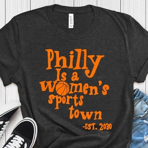 May include: Dark grey t-shirt with the text "Philly Is a Women's sports town - EST. 2030" in orange. The shirt is paired with denim shorts and black sneakers. The shirt color is H. Dark Grey.