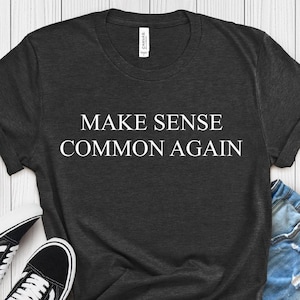 以下が含まれることがあります： 「MAKE SENSE COMMON AGAIN」の白文字がプリントされた濃いグレーのTシャツ。デニムショーツと白と黒のスニーカーを合わせています。下部のラベルには「H. DARK GREY」と書かれています。
