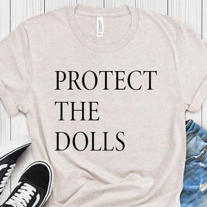 Puede incluir: Una camiseta beige claro con el texto "PROTECT THE DOLLS" impreso en negro.