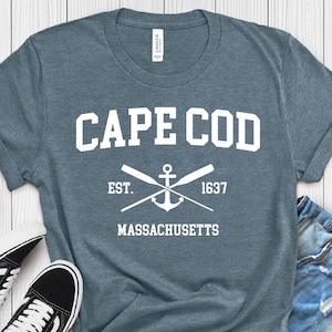 Op de afbeelding: Een heideblauw t-shirt met witte tekst die "CAPE COD EST. 1637 MASSACHUSETTS" luidt en een wit anker met gekruiste roeispanen.