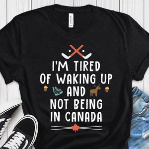 May include: Black t-shirt with the text "I'M TIRED OF WAKING UP AND NOT BEING IN CANADA" in white. The design includes hockey sticks, a maple leaf, and other Canadian icons. The shirt is labeled "VINTAGE BLACK".