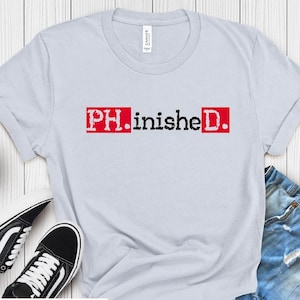 以下が含まれることがあります： 赤い背景に黒い文字で「PH.inished.」とプリントされた薄い青色のTシャツ