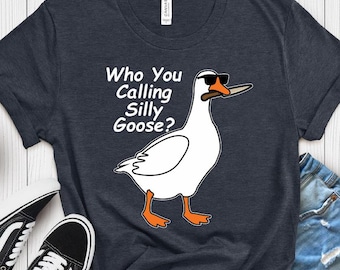 May include: A heather navy t-shirt featuring a graphic of a white goose wearing black sunglasses and holding a knife in its beak. The text "Who You Calling Silly Goose?" is printed in white next to the goose. The shirt has a crew neckline.