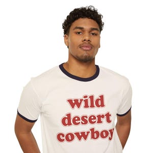 May include: A cream-coloured ringer t-shirt with navy blue trim around the collar and sleeves. The shirt displays the words "wild desert cowboy" in a bold, red, retro font. The t-shirt is made of a soft, comfortable material.