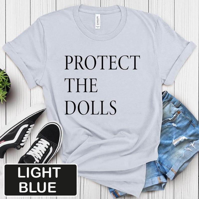 Puede incluir: Camiseta azul claro con el texto "PROTECT THE DOLLS" impreso en negro.