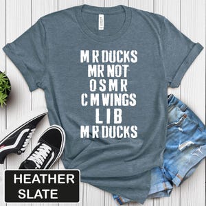 May include: Heather slate blue t-shirt with white text: "MR DUCKS MR NOT OSMR CM WINGS LIB MR DUCKS". The shirt is paired with black and white sneakers and denim shorts. The text "HEATHER SLATE" is in a black rectangle.