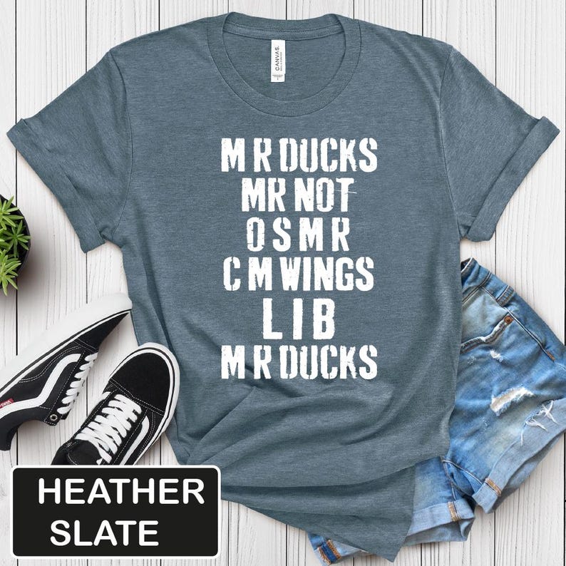 May include: Heather slate blue t-shirt with white text: "MR DUCKS MR NOT OSMR CM WINGS LIB MR DUCKS". The shirt is paired with black and white sneakers and denim shorts. The text "HEATHER SLATE" is in a black rectangle.