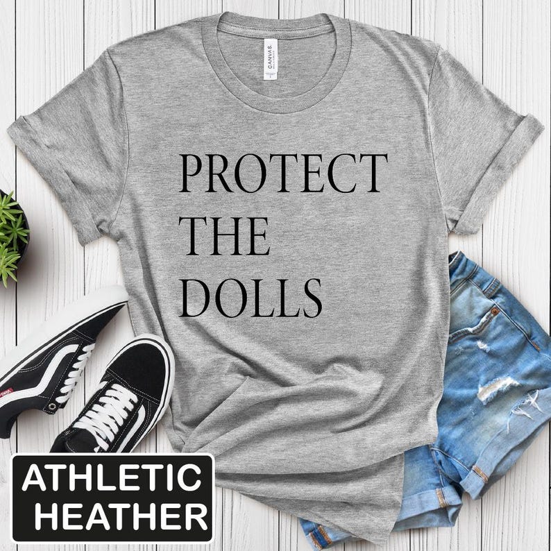 Puede incluir: Camiseta gris con el texto "PROTECT THE DOLLS" impreso en negro. La camiseta est&aacute; hecha de un material suave y c&oacute;modo y tiene un corte holgado. La camiseta se muestra sobre un fondo de madera blanco.