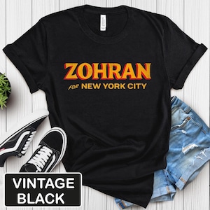 May include: Black t-shirt with the word "ZOHRAN" in yellow and red lettering, with "FOR NEW YORK CITY" below. The shirt is paired with denim shorts and black sneakers. A "VINTAGE BLACK" label is in the lower left corner.