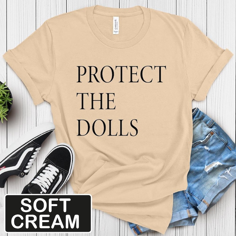 Puede incluir: Una camiseta de color crema suave con el texto "PROTECT THE DOLLS" impreso en negro.