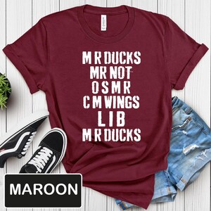 May include: Maroon t-shirt with white text that reads "MR DUCKS MR NOT OSMR CM WINGS LIB MR DUCKS." The shirt is paired with black and white sneakers and denim shorts. The word "MAROON" is in a black rectangle.