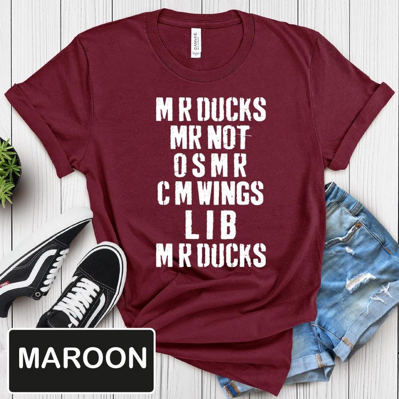 May include: Maroon t-shirt with white text that reads "MR DUCKS MR NOT OSMR CM WINGS LIB MR DUCKS." The shirt is paired with black and white sneakers and denim shorts. The word "MAROON" is in a black rectangle.