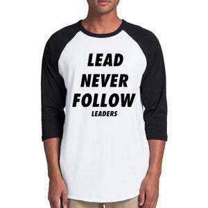 May include: A white baseball-style t-shirt with black three-quarter length sleeves and a black crew neckline. The front features the bold black text "LEAD NEVER FOLLOW" with the word "LEADERS" printed in a smaller font size directly underneath.