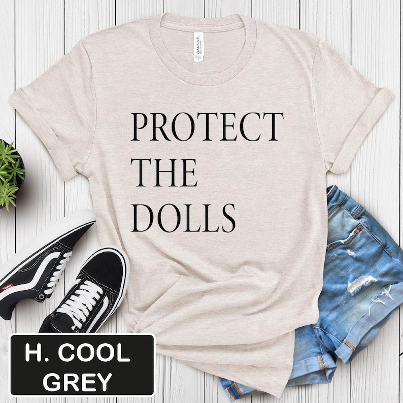 Puede incluir: Una camiseta beige claro con el texto "PROTECT THE DOLLS" impreso en negro.