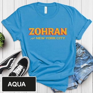 May include: Aqua blue t-shirt with the word "ZOHRAN" in orange and yellow block letters, with "FOR NEW YORK CITY" below. The shirt is displayed with black and white sneakers and denim shorts.