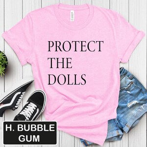 Puede incluir: Camiseta rosa con el texto "PROTECT THE DOLLS" impreso en negro.