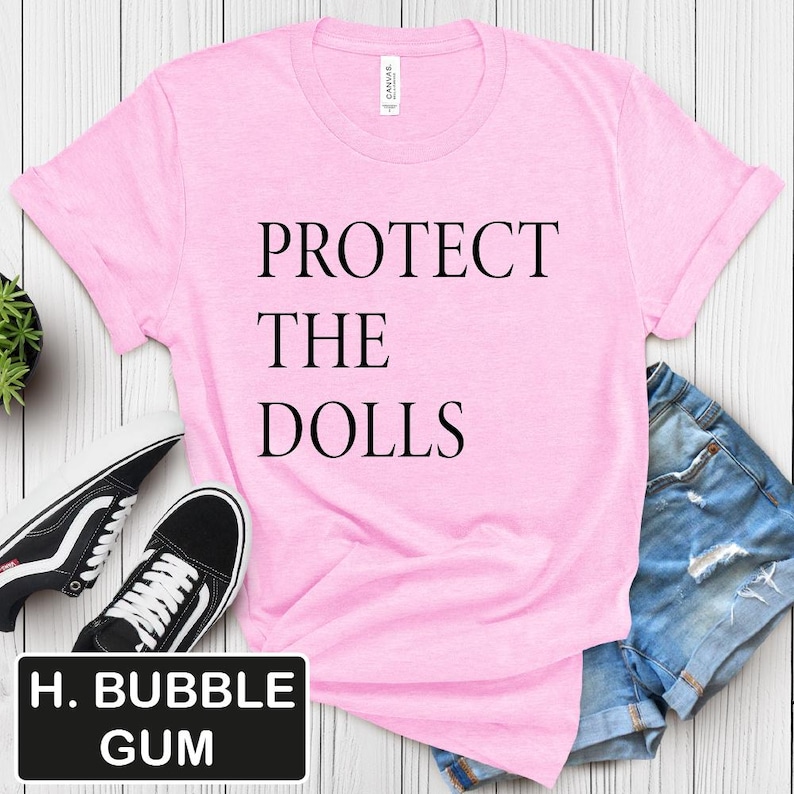 Puede incluir: Camiseta rosa con el texto "PROTECT THE DOLLS" impreso en negro.