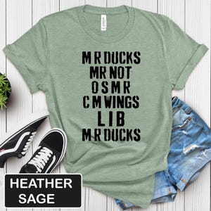 May include: Heather sage green t-shirt with the text "MR DUCKS MR NOT OSMR CM WINGS LIB MR DUCKS" in black block letters. The shirt is paired with black and white sneakers and denim shorts. The text "HEATHER SAGE" is in a black rectangle.