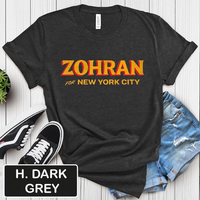 May include: Dark grey t-shirt with the word "ZOHRAN" in orange and yellow, with the text "FOR NEW YORK CITY" below. The shirt is displayed with black and white sneakers and denim shorts. The shirt color is H. DARK GREY.