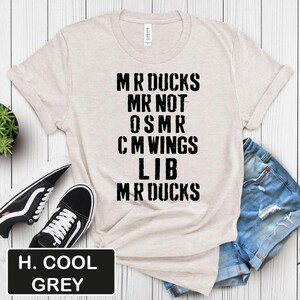 May include: A light beige t-shirt with the text "MR DUCKS MR NOT OSMR CM WINGS LIB MR DUCKS" in black. The shirt is paired with denim shorts and black and white sneakers. A black rectangle with the text "H. COOL GREY" is at the bottom.
