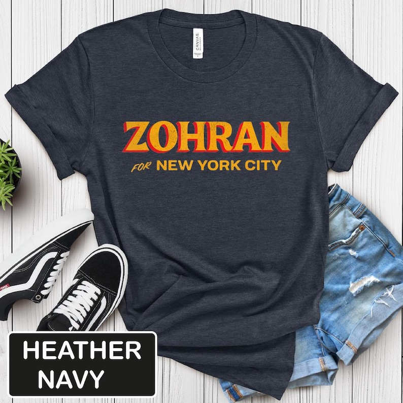 May include: Heather navy t-shirt with the text "ZOHRAN" in yellow and red, with "FOR NEW YORK CITY" below. The shirt is paired with denim shorts and black and white sneakers.