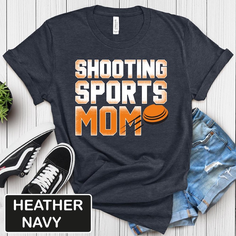 Puede incluir: Camiseta azul marino jaspeada con el texto "Shooting Sports Mom" en blanco y un gr&aacute;fico de paloma de arcilla naranja.