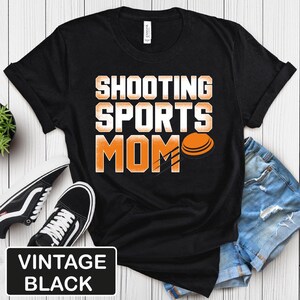 Puede incluir: Camiseta negra con el texto "Shooting Sports Mom" en letras blancas y naranjas. Se muestra un plato de arcilla junto al texto.