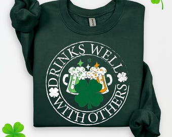 Puede incluir: Una sudadera verde oscuro con un gráfico circular blanco que dice "Drinks Well With Others" y presenta dos jarras de cerveza y un trébol.