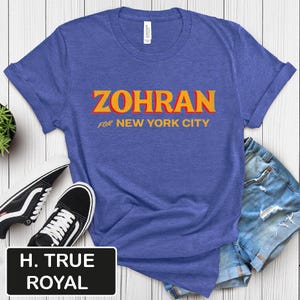 May include: A blue t-shirt with the word "ZOHRAN" in orange and yellow block letters, with "FOR NEW YORK CITY" in smaller orange letters underneath. The shirt is paired with denim shorts and black and white sneakers.