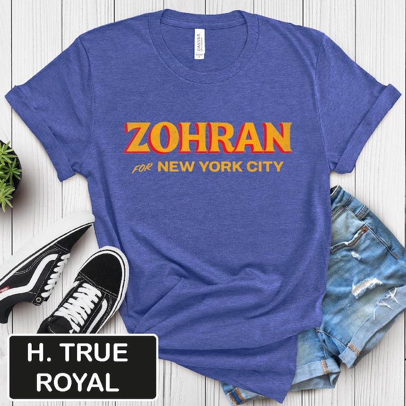 May include: A blue t-shirt with the word "ZOHRAN" in orange and yellow block letters, with "FOR NEW YORK CITY" in smaller orange letters underneath. The shirt is paired with denim shorts and black and white sneakers.