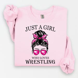 May include: Pink t-shirt with a graphic of a woman with a messy bun and sunglasses. The text "Just a girl who loves wrestling" is printed on the shirt.
