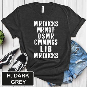 May include: Dark grey t-shirt with white text that reads "MR DUCKS MR NOT OSMR CM WINGS LIB MR DUCKS." The shirt is paired with denim shorts and black and white sneakers. The bottom left corner of the image has a black rectangle with the text "H. DARK GREY."
