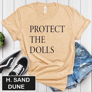 Puede incluir: Una camiseta beige claro con el texto "PROTECT THE DOLLS" impreso en negro. La camiseta est&aacute; sobre una superficie de madera con un par de zapatillas negras y unos pantalones cortos de mezclilla azul.