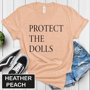 Puede incluir: Una camiseta de color melocot&oacute;n brezo con el texto "PROTECT THE DOLLS" impreso en negro.
