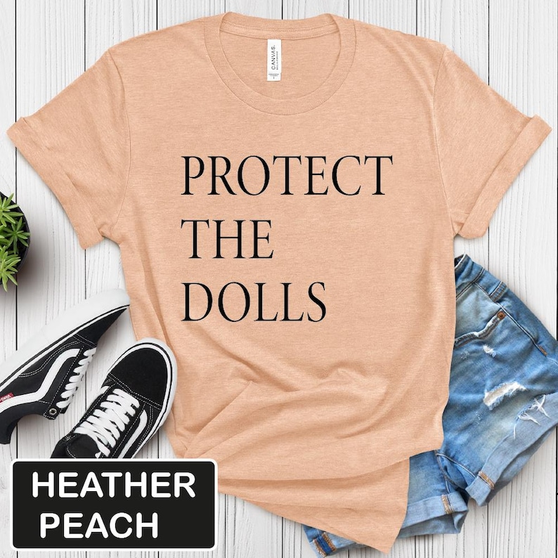 Puede incluir: Una camiseta de color melocot&oacute;n brezo con el texto "PROTECT THE DOLLS" impreso en negro.