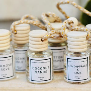 May include: A collection of small glass diffuser bottles with wooden tops and hanging cords. Each bottle has a label with a scent name, including "Oakmoss Grove" and "Coconut Sands". The bottles are arranged on a light-colored surface.