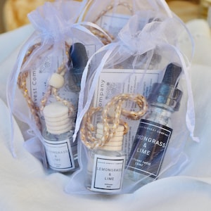 May include: Three small glass bottles of lemongrass and lime diffuser refills are presented in sheer white bags. Each bottle has a wooden top and a label with the text "Lemongrass & Lime". The bags are tied with gold and white cords.