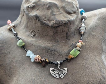 May include: A necklace featuring a silver-toned ginkgo leaf pendant centered on a strand of black beads interspersed with various multi-colored stones, including green, blue, brown, and patterned beads in assorted shapes and sizes.