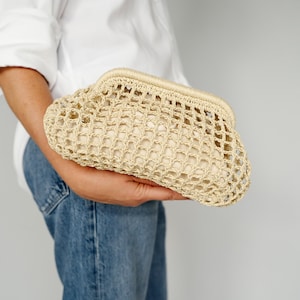 May include: A beige, woven clutch with a net-like design. The clutch is held in the hand and has a rounded shape with a structured frame. The bag is made of a natural fiber material.