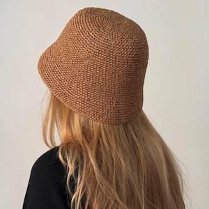 May include: A brown woven bucket hat with a rounded crown and a slightly downward-sloping brim. The hat is made of a textured material. The hat is worn by a person with long, blonde hair and a black top. A stylish accessory.